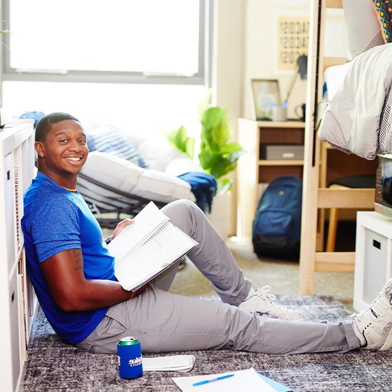 A Drake University student sitting on the floor of a dorm room holding a notebook and smiling